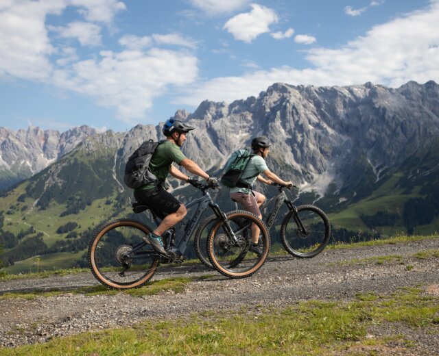 medium-Paerchen-geniesst-Tour-mit-dem-eBike-vor-herrlichem-Bergpanorama Dva cyklisté jedou na elektrokole Hochkönig po štěrkové cestě s malebnými horami a zelenými údolími v pozadí.