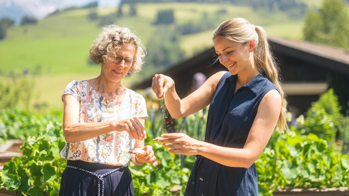 Dvě ženy se usmívají a připravují jídlo s bylinky Hochkönig ve slunné zahradě s kopci v pozadí.