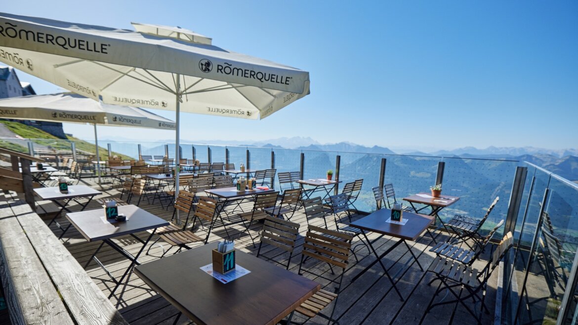 Na vrcholu Schafbergu čeká občerstvení s impozantním výhledem © Salzburg AG Tourismus GmbH Venkovní terasa s prázdnými stoly, výhledem na lanovku Zubačka Schafbergbahn a za jasného počasí s velkými bílými slunečníky. (Zvětšený pohled)