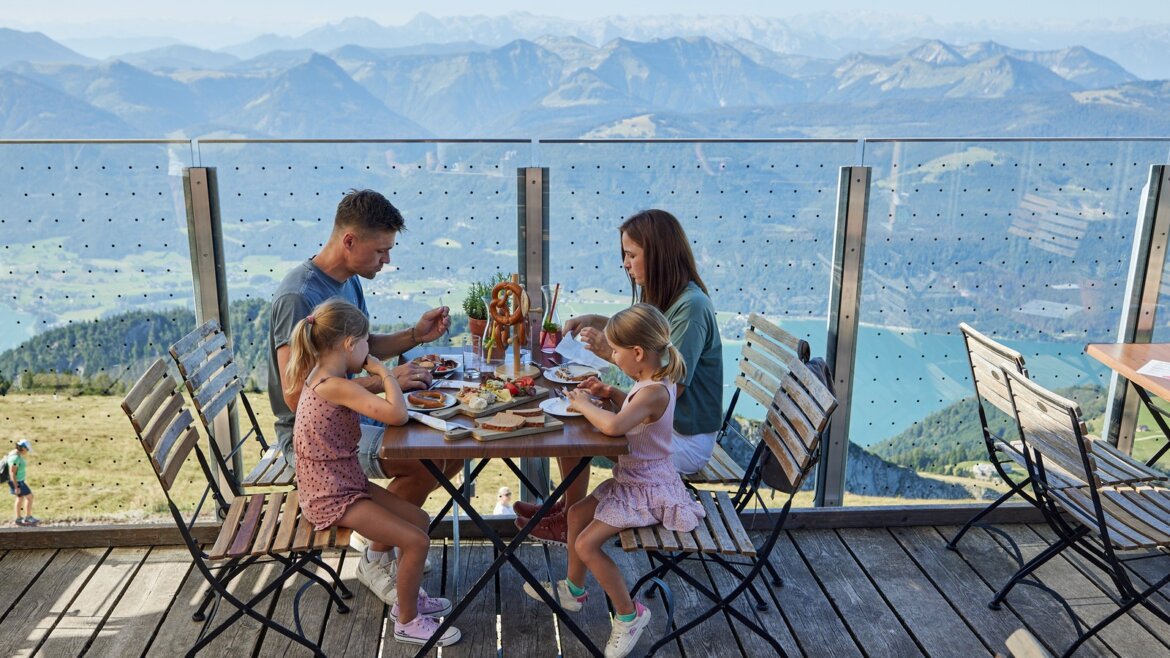 Výletní restaurace na vrcholu Schafbergu © Salzburg AG Tourismus GmbH Čtyřčlenná rodina obědvá pod širým nebem s výhledem na jezera a vrcholy v blízkosti malebných horských vyhlídek lanovky Zubačka Schafbergbahn. (Zvětšený pohled)