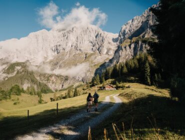 Dva turisté jdou po stezkách směrem k chatě, obklopeni trávou, stromy a vrcholy Turistika Hochkönig.