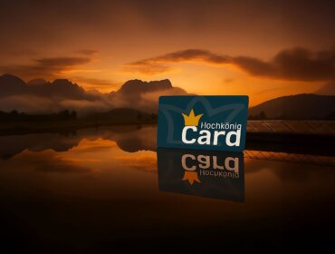 Karta Hochkönig Card stojí na zrcadlícím se jezeře při západu slunce, v pozadí jsou hory.