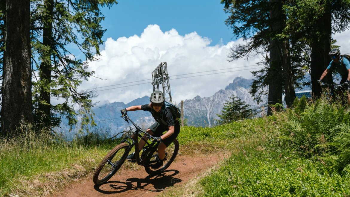 Horský cyklista v helmě jede po flowtrailu na sjezdovce Hochkönig, v pozadí jsou hory a mraky.