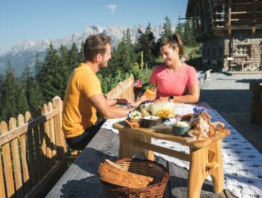 Pár si vychutnává na terase s výhledem na hory tradiční alpskou svačinu složenou z domácích dobrot, špeku, uzenin, sýrů, pomazánek, zeleniny a bylinek