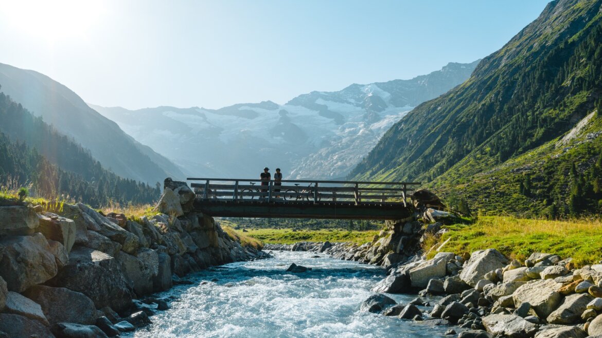 Dva lidé stojí na dřevěném mostě s Elektrokolou Vysoké Taury, vrcholy a lesem v pozadí.