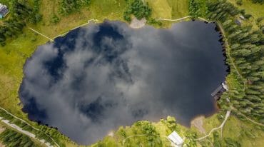 Preberské jezero Prebersee vyfocené z ptačí perspektivy. V temné hladině se odráží nebe, všude okolo zelené koruny stromů