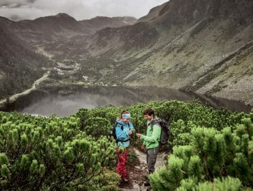 Dva turisté stojí na zelené horské stezce s výhledem na jezero a údolí obklopené kopci.