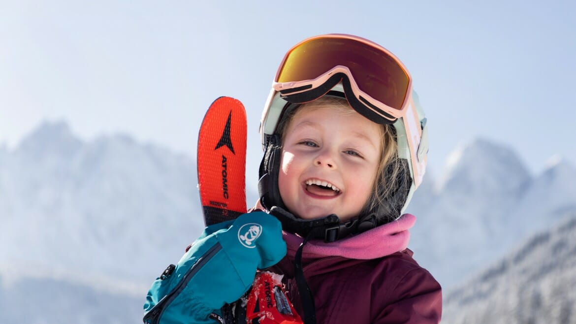 Rozesmáté dítě s lyžemi, helmou a lyžařskými brýlemi na sjezdovce v rodinné oblasti Dachstein West