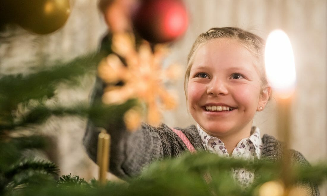 Dívka věší červenou vánoční ozdobu na stromek, na kterém už je i zlatá svíčka a slaměná hvězdička