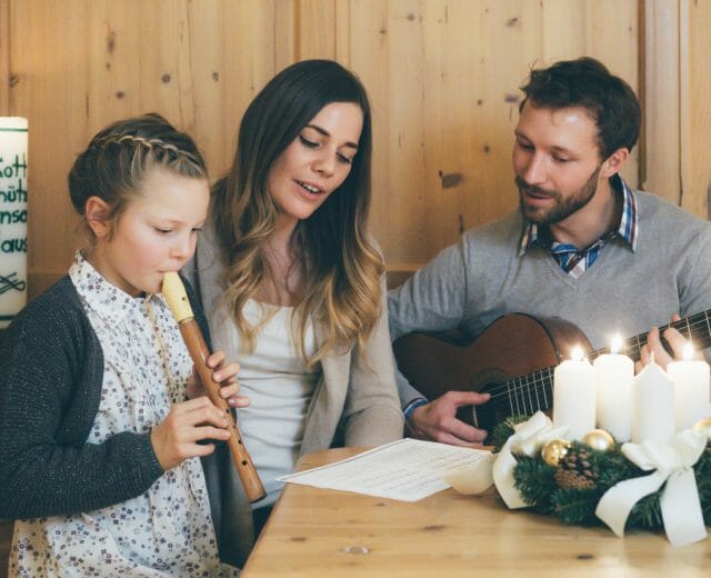 Familie musiziert im Advent Tatínek na kytaru, maminka zpívá, dcera hraje na flétnu u stolu s adventním věncem koledu Tichá noc, svatá noc.