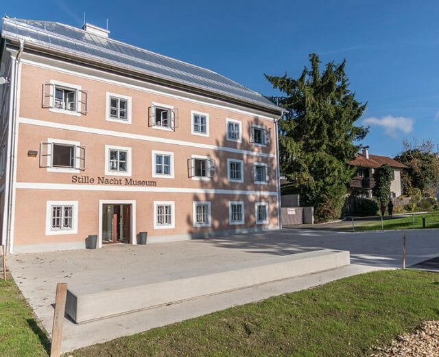 Třípatrová růžová budova s nápisem Muzeum Tiché noci Oberndorf a zeleným trávníkem pod modrou oblohou.