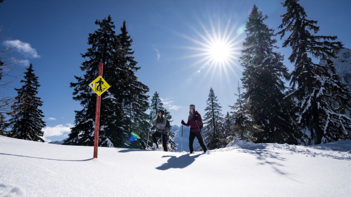 Dva lidé procházejí hlubokým sněhem kolem žluté značky pro turistiku na sněžnicích v Dachstein West. 
