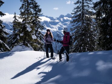 Dva lidé stojí v hlubokém sněhu na výpravě na sněžnicích v Dachstein West, za nimi zasněžené stromy a vrcholy.