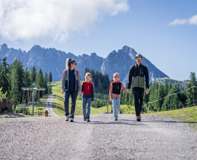 BrunosBergwelt – Familienerlebnis (c) DAG, ChrisEderMedia Čtyřčlenná rodina jde společně po štěrkové cestě během rodinné dovolené v Dachstein West.