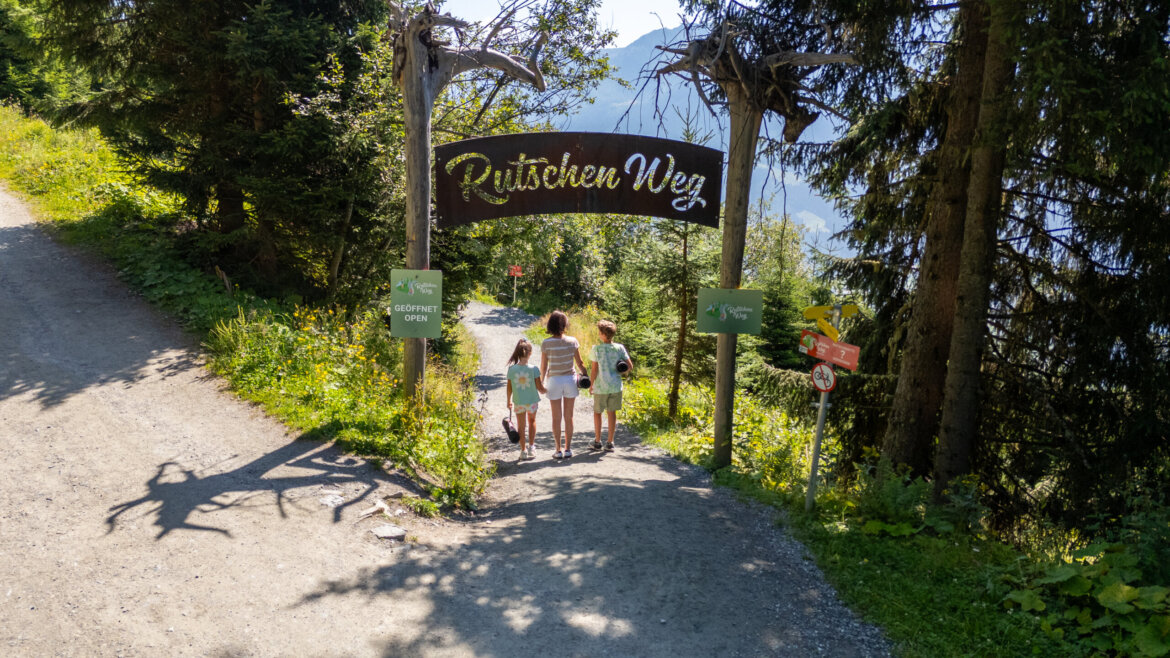 Tři děti se procházejí pod dřevěnou cedulí Rutschen Weg ve slunné Léto Wildkogel-Areně Neukirchen Bramberg.