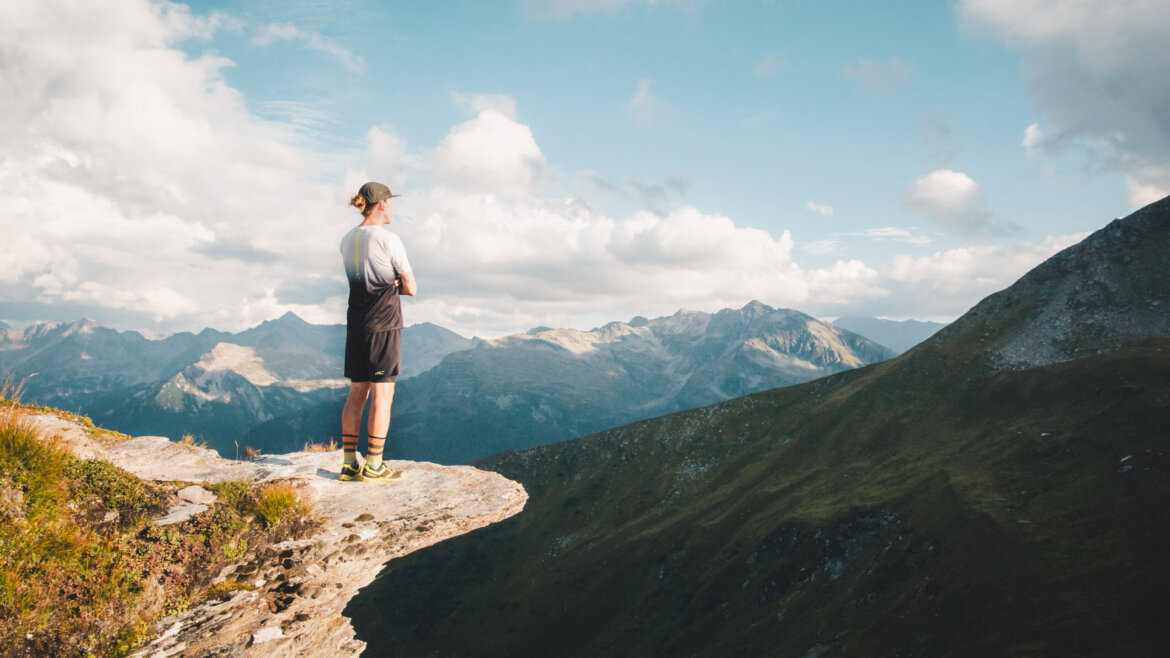 Osoba stojící na okraji skalního útesu s výhledem na vzdálené pohoří na 16. etapě dálkové trasy Hohe Tauern Panorama Trail (Zvětšený pohled)