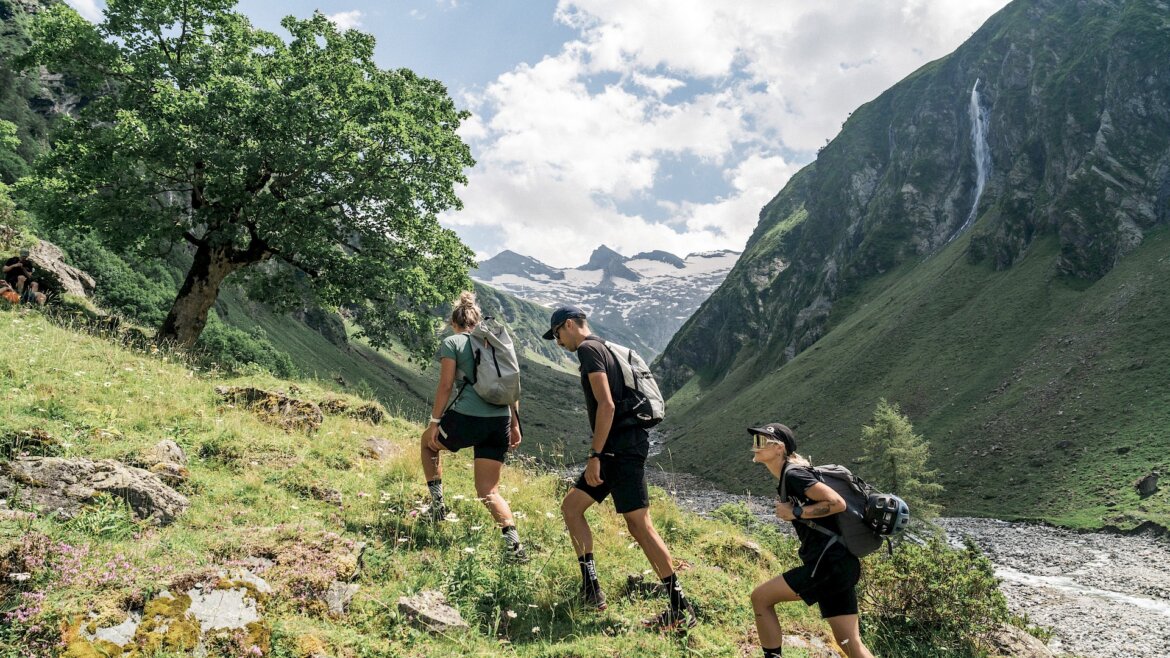 Tři lidé na túře v Léto Wildkogel-Arena Neukirchen Bramberg, za nimi strom, vodopád a zasněžené vrcholky.