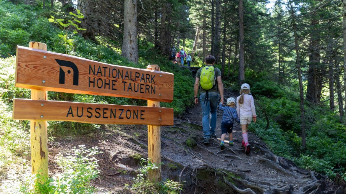 Rodina jde po lesní stezce kolem cedule Nationalpark Hohe Tauern v Raurisertalu 