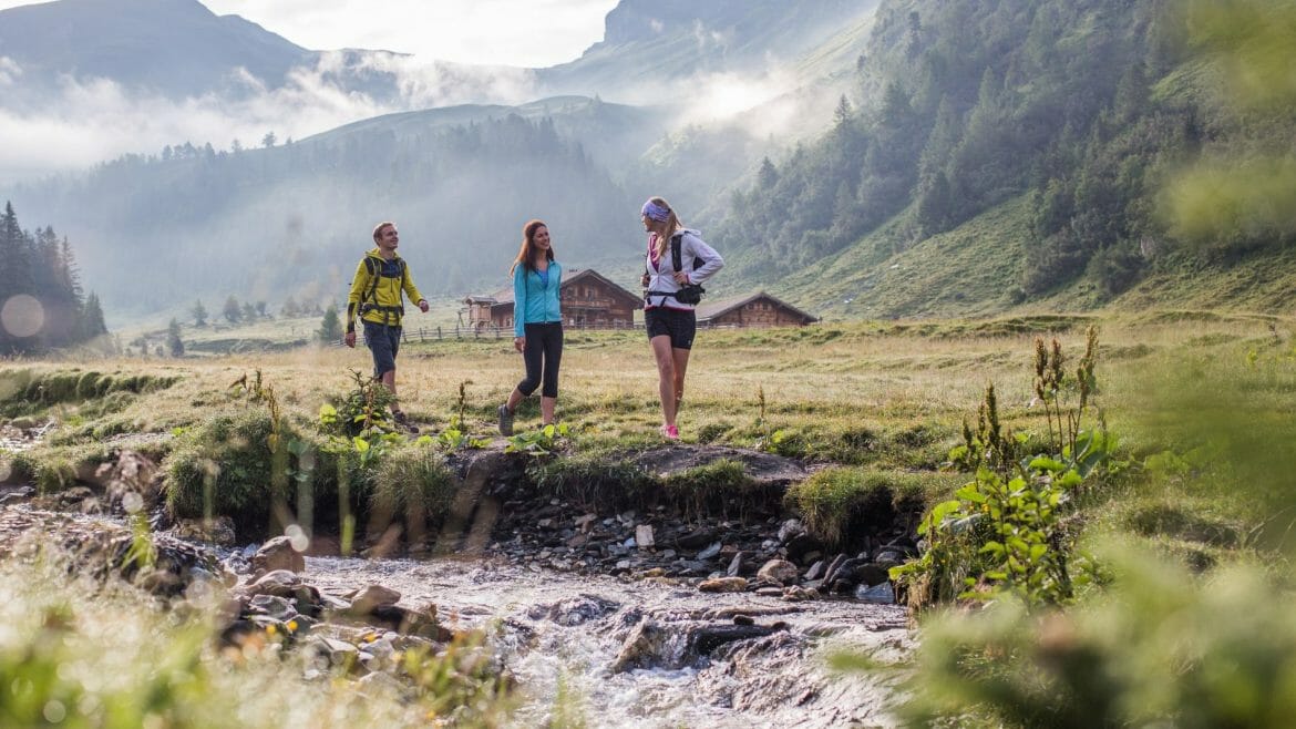 Tři turisté putují podél potoka v Raurisertalu mezi svěžími zelenými kopci 