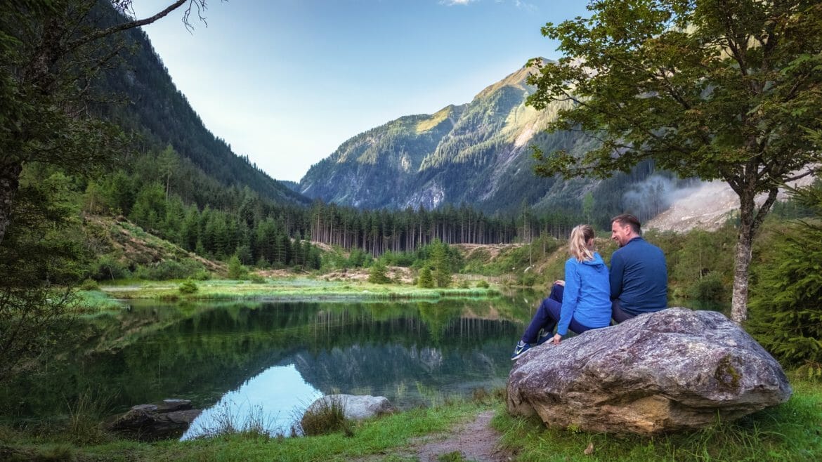 Pár odpočívá na kameni u jezera Blausee s tyrkysově modrou vodou v chráněné krajinné oblasti, kde voní smrky, jedle a modříny.
