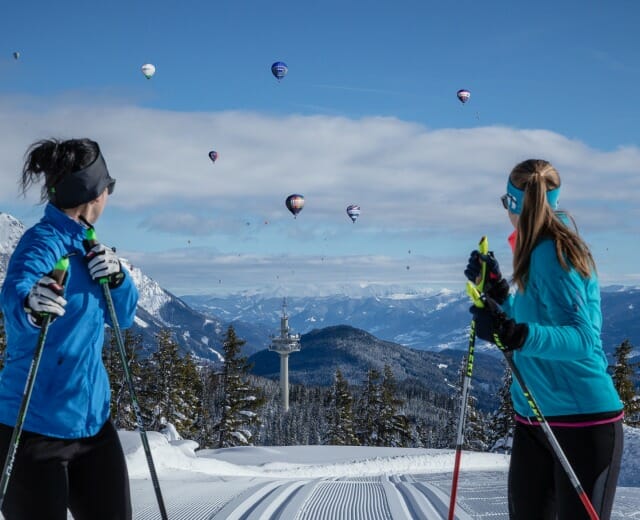 Ballon und Ski am Rossbrand in Filzmoos©CoenWeesjes_www.filzmoos.at Běžkařky pozorují z vysokohorské běžecké tratě na hoře Rossbrand v oblacích balóny, které vzlétají z Filzmoosu