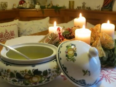 Štědrovečerní vývar z hovězího masa se zeleninou, uzeninami a nudlemi servírovaný v malované porcelánové míse, zatímco na stole hoří na adventním věnci všechny čtyři svíčky.