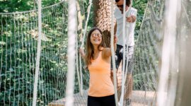 21_st_johann_in_salzburg_sommer_baumparcours_klettern_3 Dva lidé přecházejí přes lanový most v lese, užívají si sportovní aktivity v St. Johann v Salcbursku.