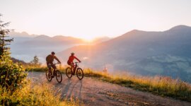 Dva lidé jedou na horských kolech při západu slunce v malebném St. Johann na kole s kopci a údolími v pozadí.