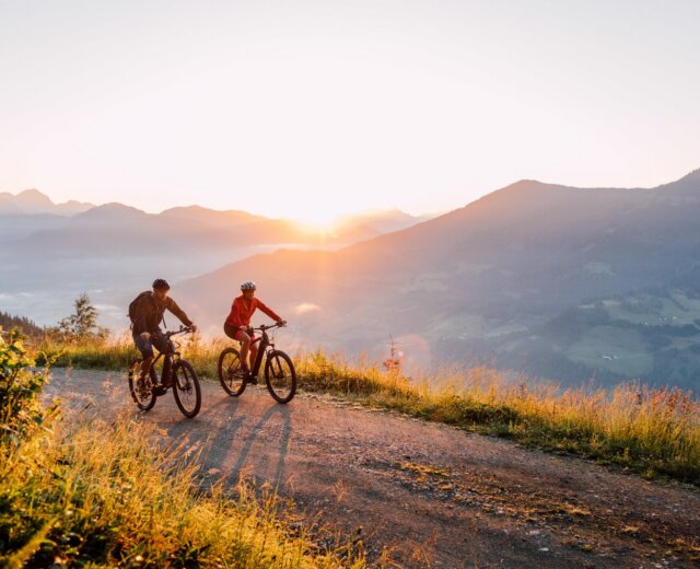 23_st_johann_in_salzburg_sommer_mountainbike_sonnenaufgang_8 Dva lidé jedou na horských kolech při západu slunce v malebném St. Johann na kole s kopci a údolími v pozadí.
