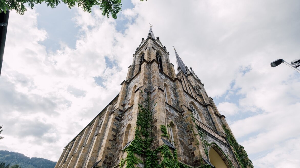 Pohled na Pongauský dóm v obci St. Johann v Salcbursku s břečťanem na stěnách a věžích pod zataženou oblohou. (Zvětšený pohled)