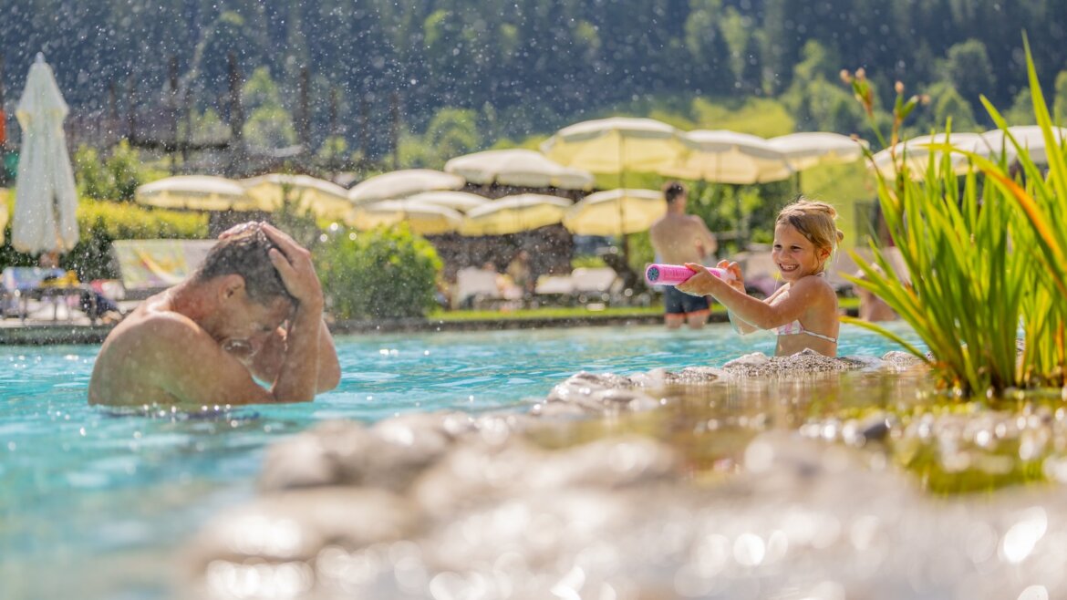 Usmívající se dítě stříká vodu na muže ve slunném bazénu ve Flachau na Salcbursku, za nímž jsou slunečníky a stromy. (Zvětšený pohled)
