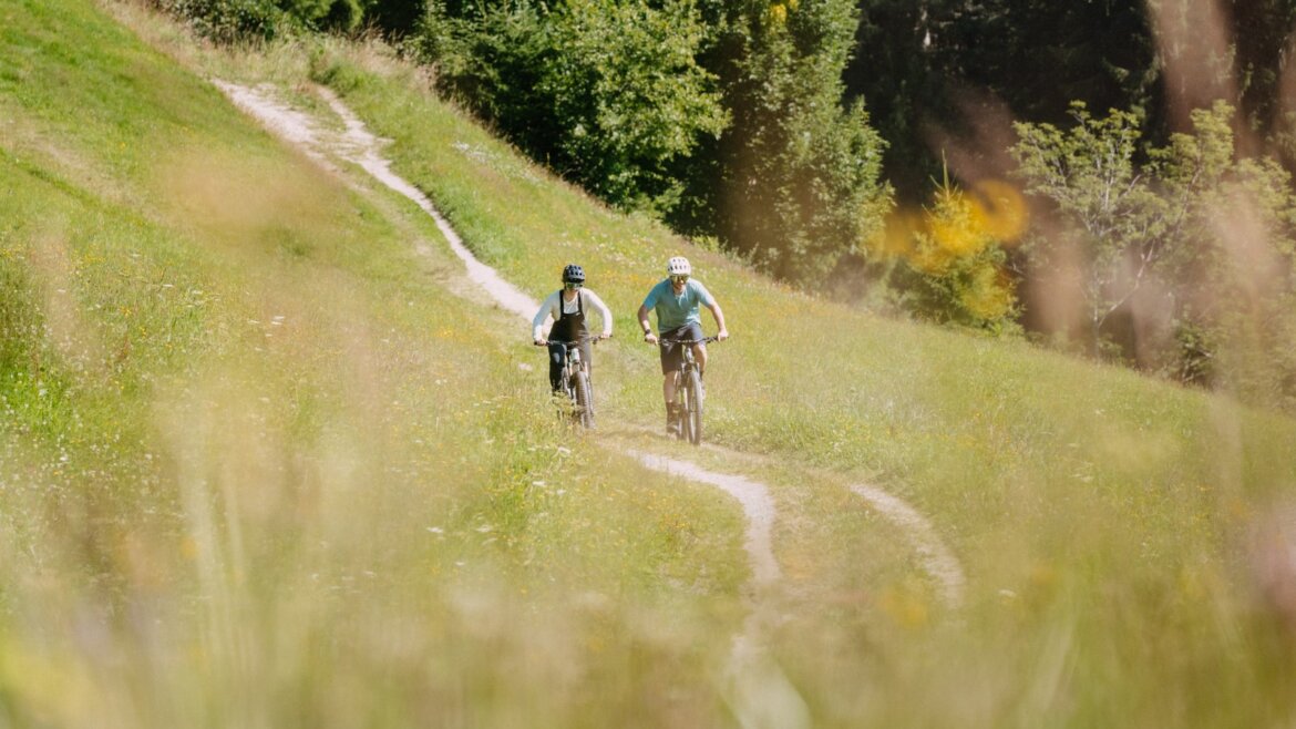 Dva lidé jedou na kolech po klikaté stezce přes travnaté pole a na slunci popíjejí flachau kolu.