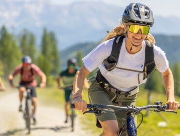Skupina cyklistů jede na horských kolech po prosluněné stezce Flachau kola se stromy a horami v pozadí.