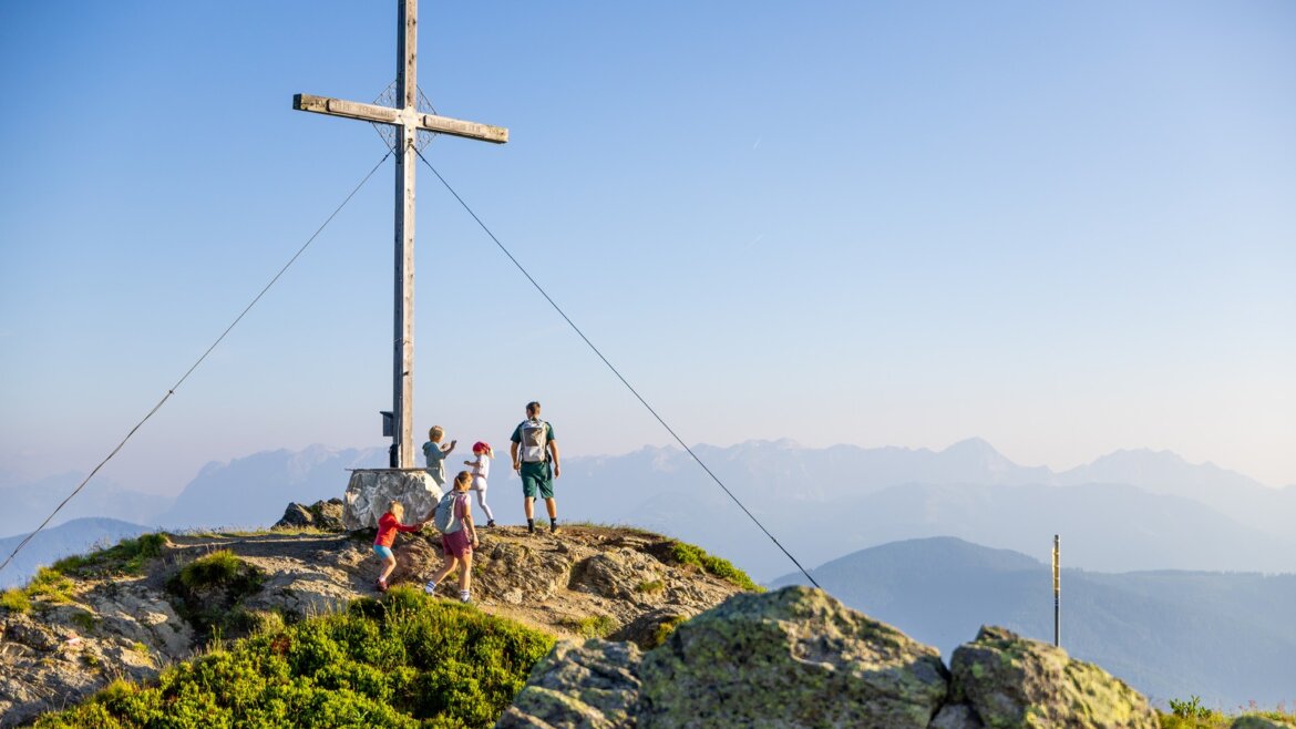 Hlavní flachauská hora Grießenkar © Flachau Tourismus, Markus Berger Čtyři lidé stojí u velkého dřevěného kříže na horském vrcholu Flachau turistika s výhledem na vzdálené kopce. (Zvětšený pohled)