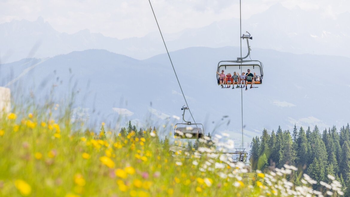 Jízda lanovkou je pro děti zážitek © Flachau Tourismus, Markus Berger Lidé jedoucí na lyžařském vleku nad rozkvetlou loukou ve Flachau turistika, s horami a stromy v pozadí. (Zvětšený pohled)
