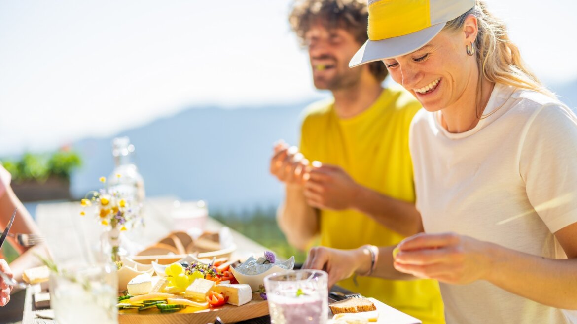 Dvě osoby si vychutnávají slunečné jídlo, dělí se o občerstvení a nápoje Flachau kuchyně u dřevěného stolu pod širým nebem. (Zvětšený pohled)