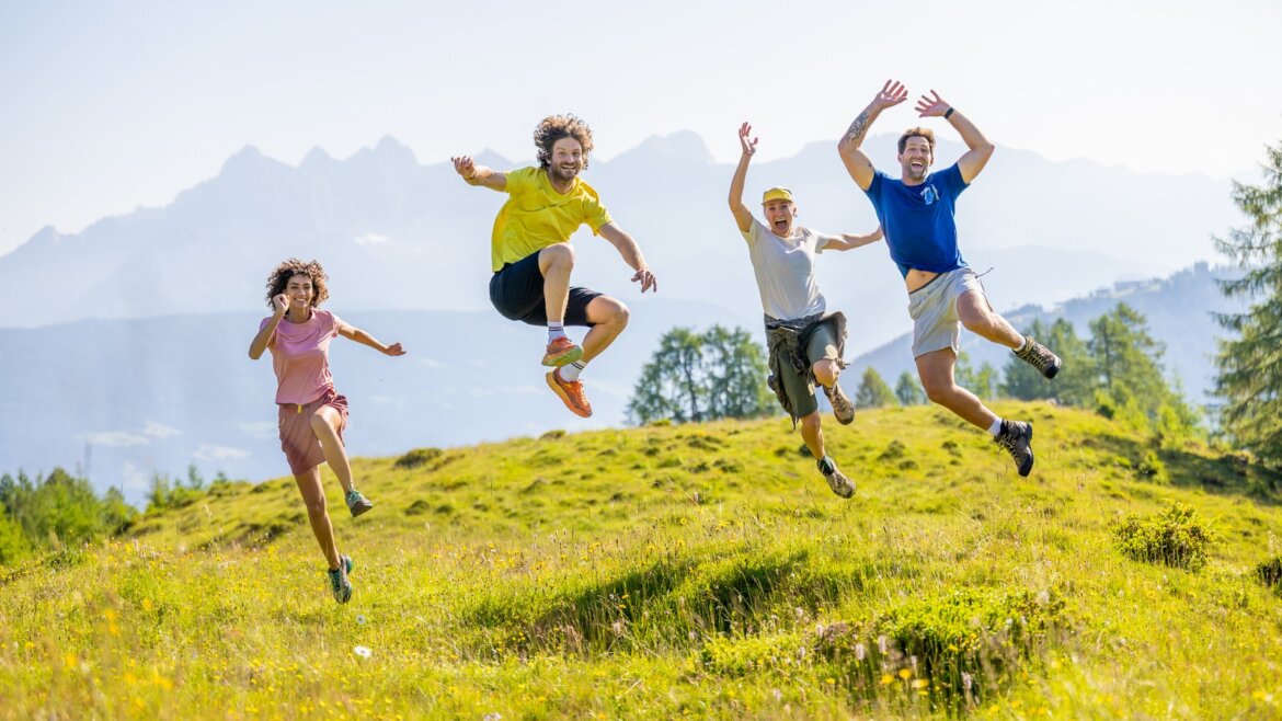 Čtyři lidé radostně skáčou na travnaté louce Flachau turistika s horami a stromy v pozadí.