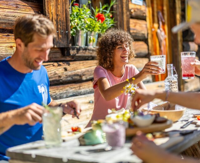Tři lidé se usmívají a společně si připíjejí nápoji u rustikálního stolu a užívají si Flachau kuchyně pod širým nebem.