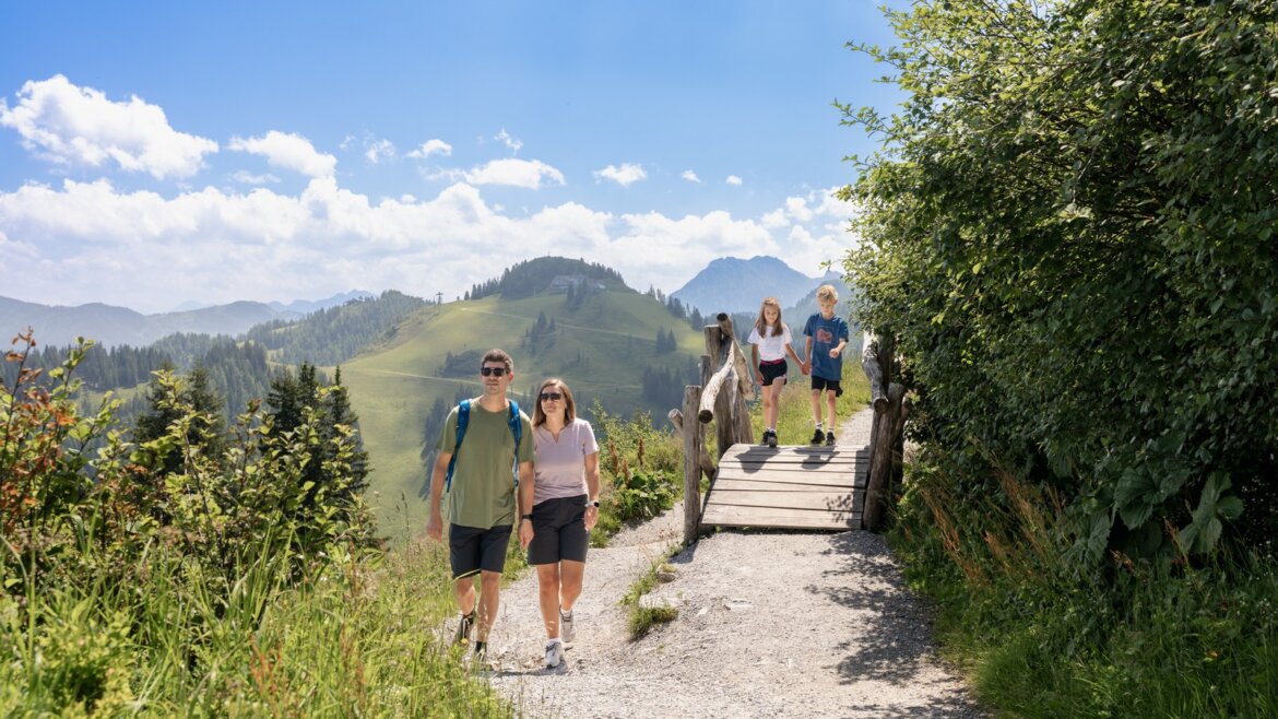 Rodinný výlet na Geisterbergu © JOsalzburg Čtyři lidé se vydávají po slunné horské stezce na rodinnou dovolenou Svatý Jan léto, přechází malý dřevěný most. (Zvětšený pohled)