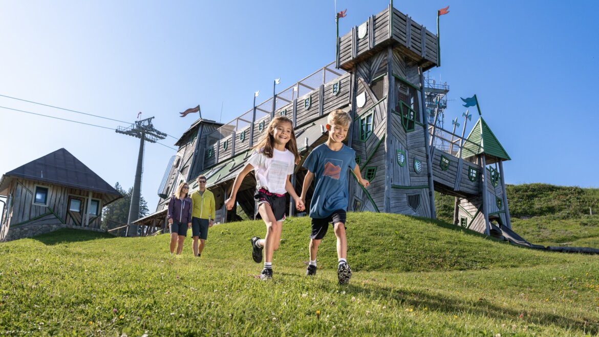 Dvě děti běhají po trávě a dospělí je pozorují, jak si užívají Rodinná dovolená sv. Jana pod jasnou modrou oblohou.