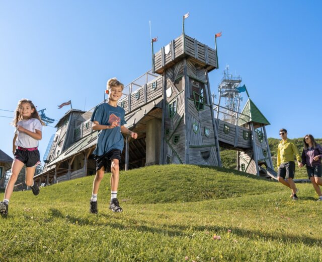 Dvě děti běží z travnatého kopce u Letních lanovek Svatý Jan v Salcbursku pod jasnou modrou oblohou.