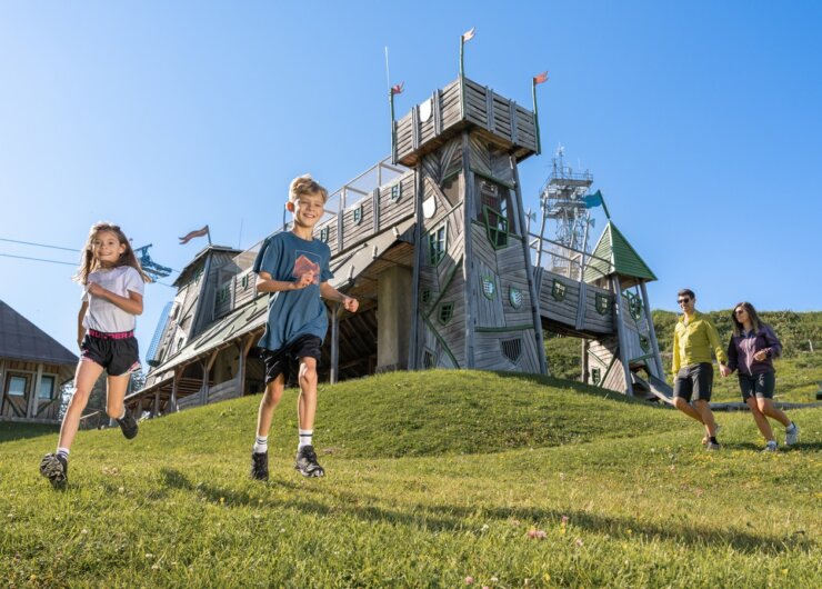 25_st_johann_in_salzburg_sommer_geisterberg_familie_27 Dvě děti běží z travnatého kopce u Letních lanovek Svatý Jan v Salcbursku pod jasnou modrou oblohou.