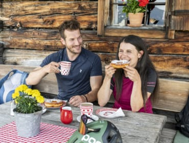 almjause-in-altenmarkt-zauchensee Muž a žena sedí před rustikální chatou a vychutnávají si Altenmarkt-Zauchensee kuchyně s kávou a pečivem.