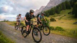 Tři lidé na Königstour s přepravou zavazadel, jízda na kole po šotolinové stezce přes zelené kopce a horské výhledy.