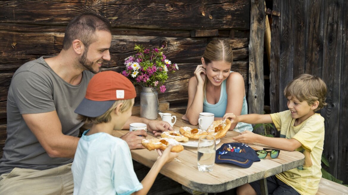 Čtyřčlenná rodina si pochutnává na pečivu u rustikálního srubu, který evokuje kouzlo Kuchyně ve Svatém Janu na Salcbursku. (Zvětšený pohled)