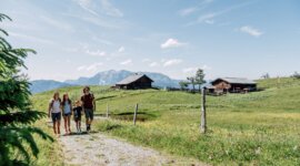 Čtyři lidé jdou po travnaté cestě poblíž turistických chatek St. Johann s horami a modrou oblohou za zády.