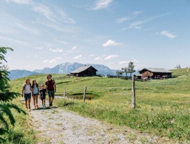 Čtyři lidé jdou po travnaté cestě poblíž turistických chatek St. Johann s horami a modrou oblohou za zády.