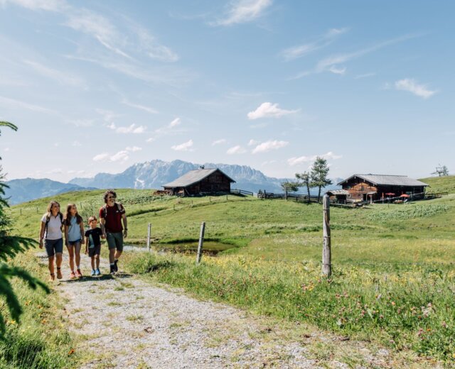 Čtyři lidé jdou po travnaté cestě poblíž turistických chatek St. Johann s horami a modrou oblohou za zády.