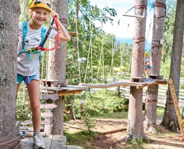 hochseilgarten-in-altenmarkt-zauchensee-1170×658 Dítě s helmou a jištěním prochází mezi stromy na stezce v lanovém parku v Altenmakrt-Zauchensee.