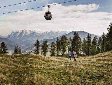 Dva turisté se procházejí po travnaté alpské louce, zatímco jim nad hlavou kloužou Letní lanovky sv. Jana v Salcbursku.